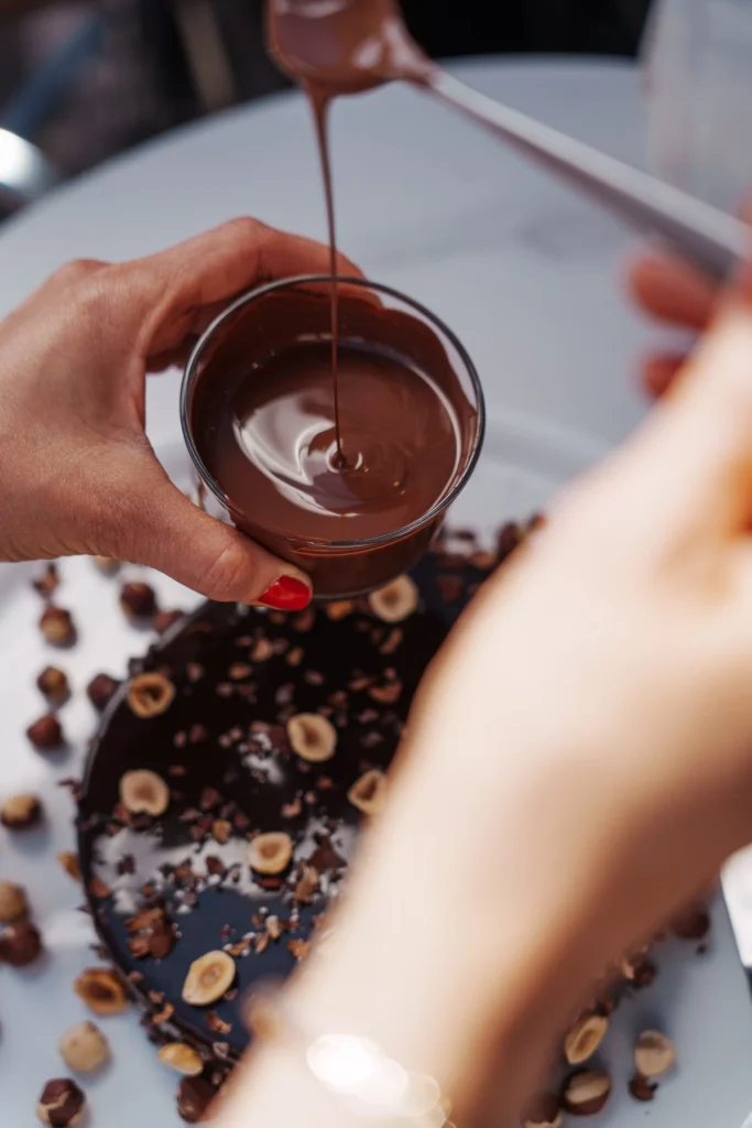 Une main tient un bol de chocolat fondu au-dessus d’un gâteau décoré de noisettes chez Graine de Vanille